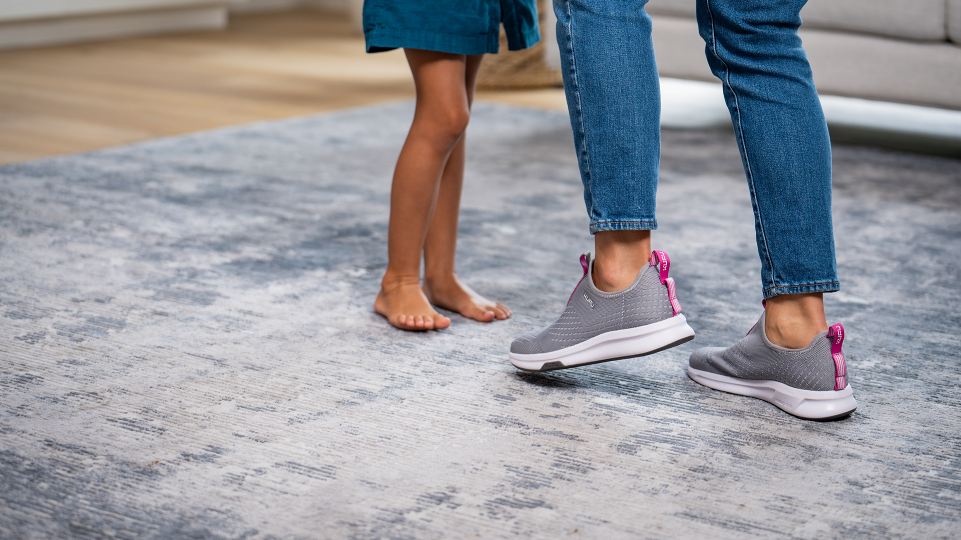 A person with their child, focusing on the feet of each. The mother wears comfortable KURU Footwear shoes on a fluffy rug